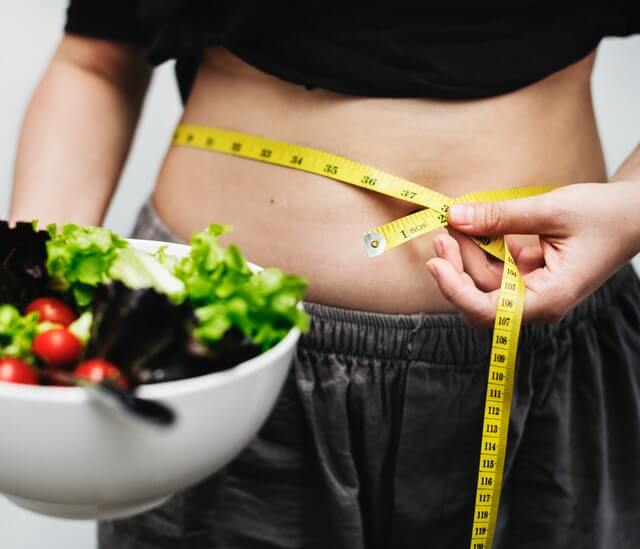 A imagem mostra uma mulher medindo sua barriga com fita métrica e um bowl de salada