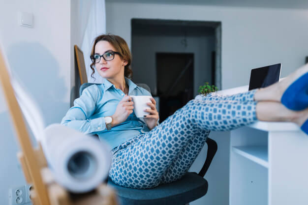 mulher de óculos sentada na cadeira tomando um café com as pernas em cima da mesa relaxando