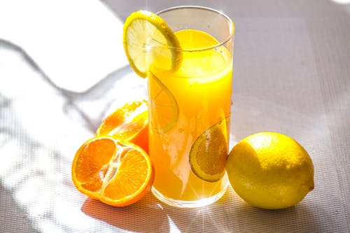 Copo de suco de laranja em cima da mesa, com uma laranja inteira e uma laranja cortada ao meio; no copo, uma rodela da fruta.