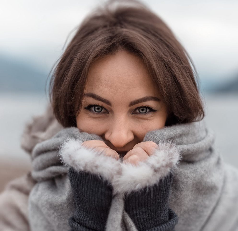 Na imagem, há uma mulher com roupas de inverno, com as mãos próximas ao rosto, cobrindo-o até a altura do nariz