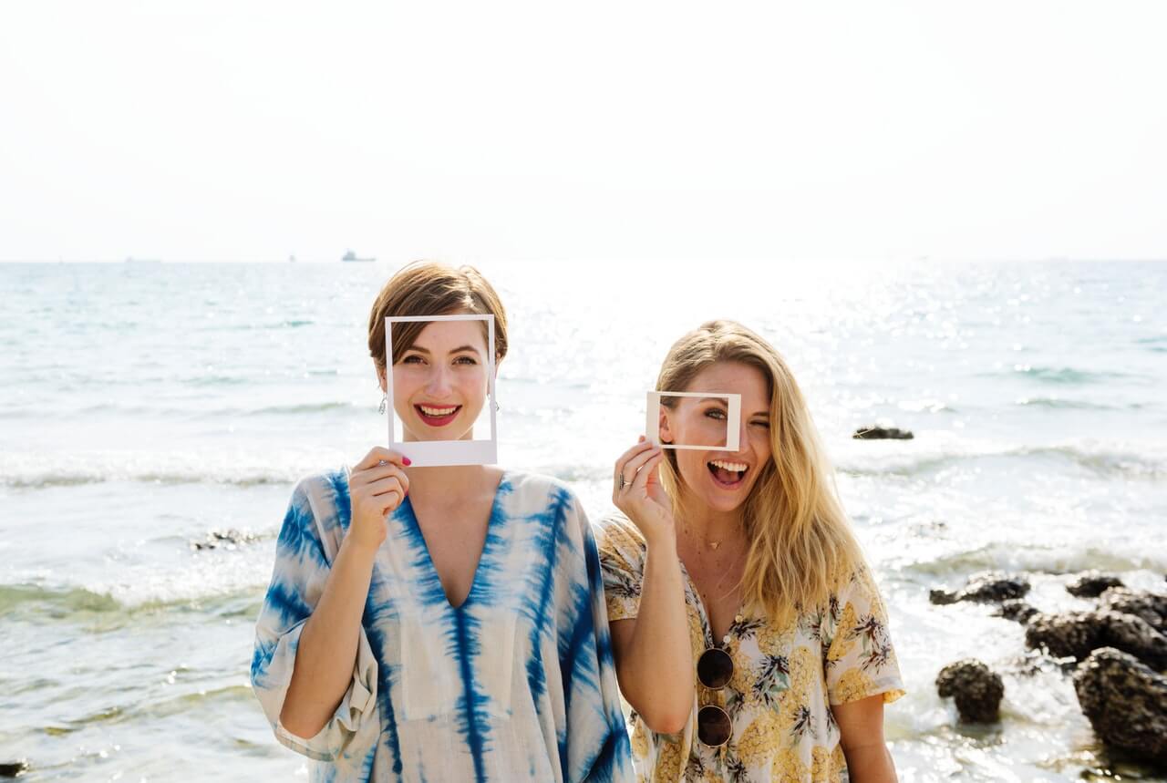 Duas mulheres posando para a foto na praia, segurando molduras pequenas