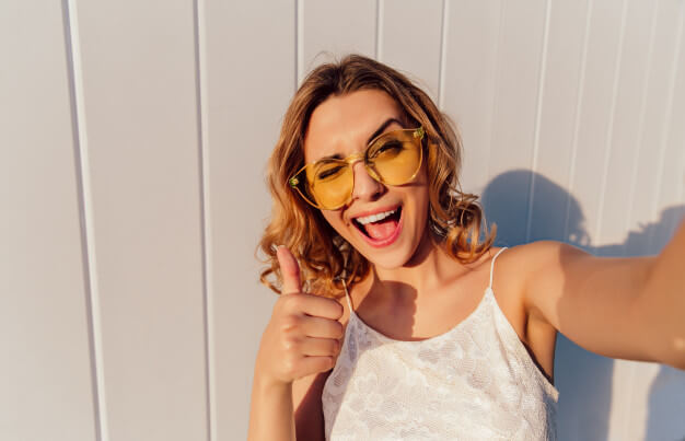 mulher tirando uma selfie de óculos amarelo sorrindo e fazendo sinal de positivo